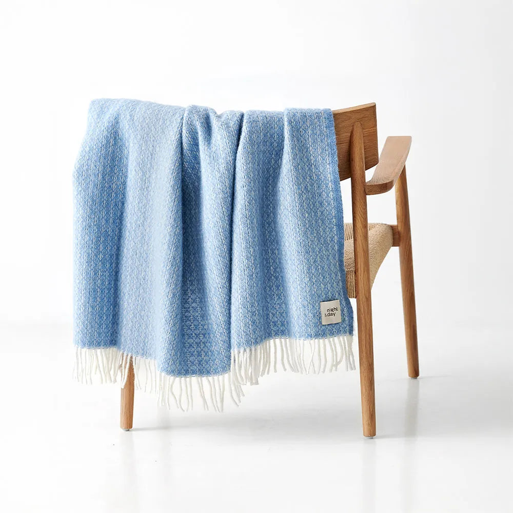 Lyseblå uldplaid fra Night & Day hænger over en træstol med hvid baggrund – WOOLMARK-certificeret 100% uld