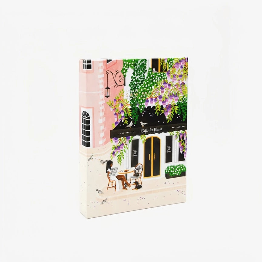Paris-puslespil fra All the Ways to Say med motiv af en fransk café omgivet af grønne planter og lilla blomster.