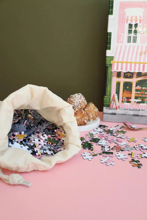 Pink Bakery-puslespilsbrikker i en stofpose og på et rosa bord med æsken i baggrunden