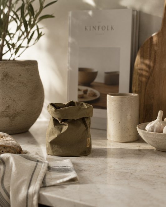 Uashmama pose small i oliven på marmorbord med oliventræ, keramik og Kinfolk-bog i baggrunden
