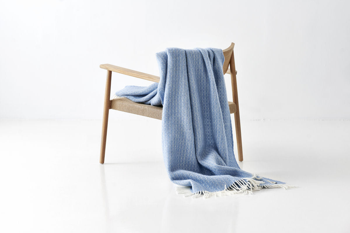 Lyseblå uldplaid fra Night & Day hænger over en træstol med hvid baggrund – WOOLMARK-certificeret 100% uld