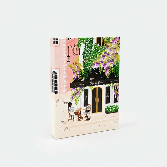 Paris-puslespil fra All the Ways to Say med motiv af en fransk café omgivet af grønne planter og lilla blomster.